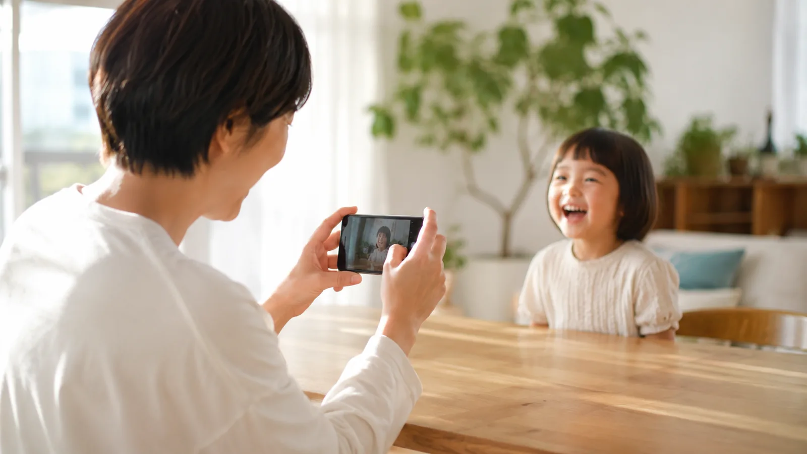 明るいリビングでスマートフォンを使い、笑顔の子どもを撮影する親
