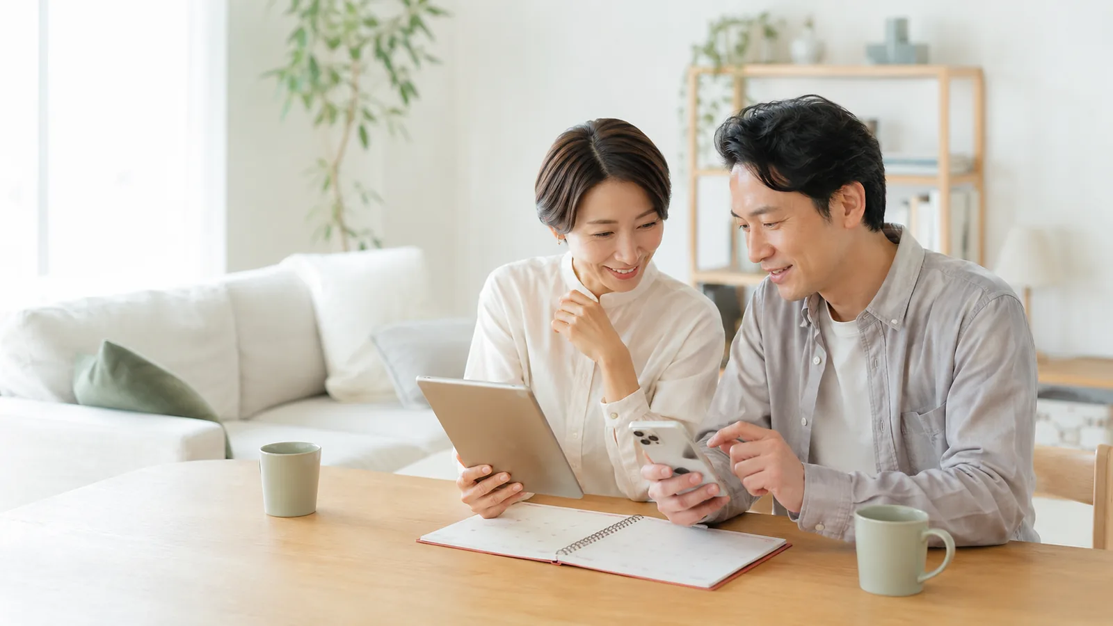 明るいリビングで夫婦がスマホとカレンダーを見ながら、予定や情報を共有している様子